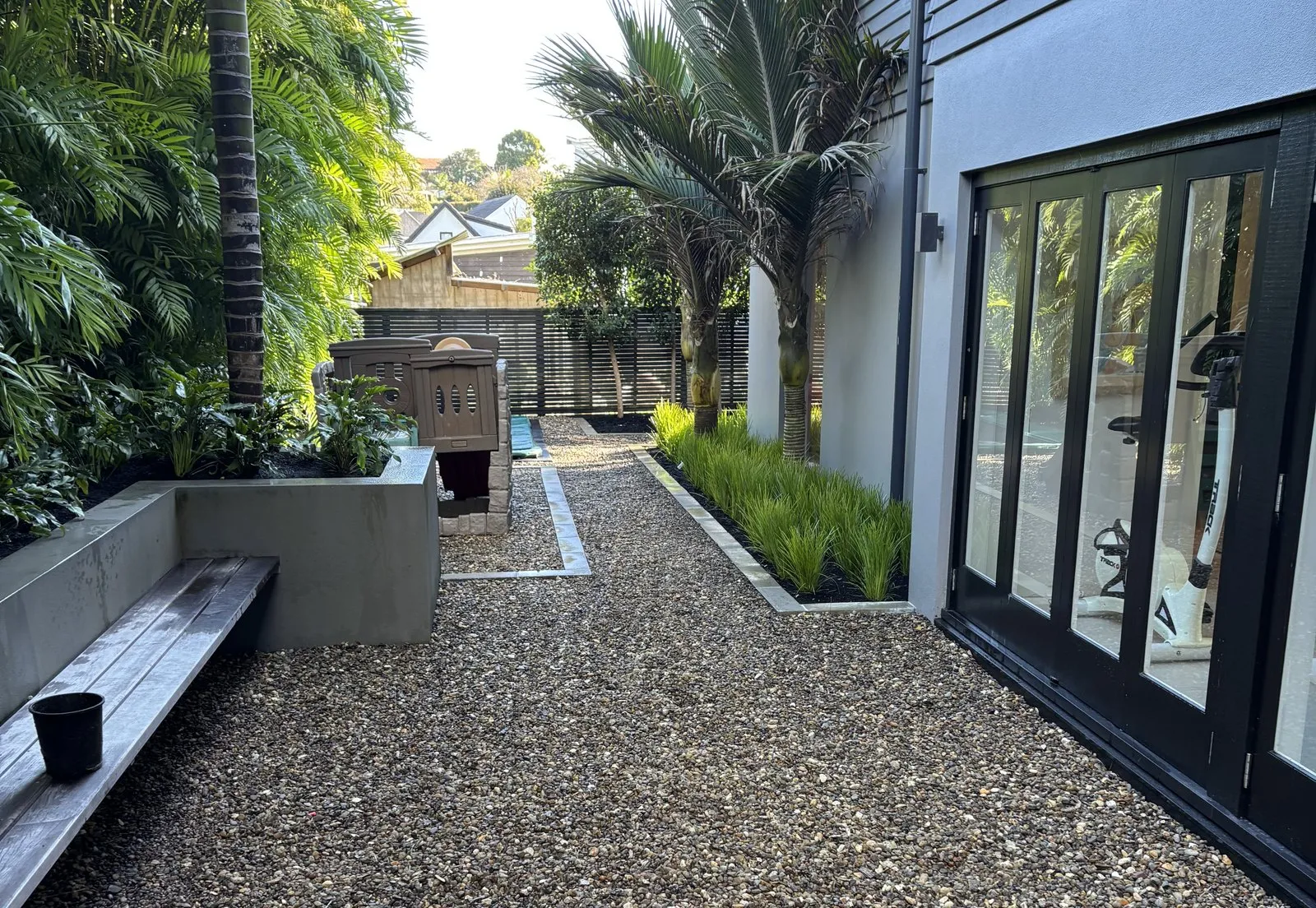 A gravel courtyard beside a modern building, with palms, a planter bench and a grass border